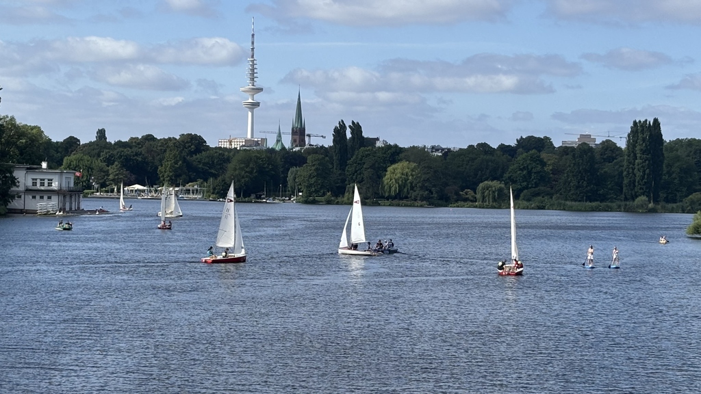 Segelboote auf der Alster mit Uferbäumen; Ferne Stadt skyline mit Turm und Kirchtürmen.